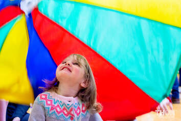 Image: child smiling and looking up under a rainbow parachute